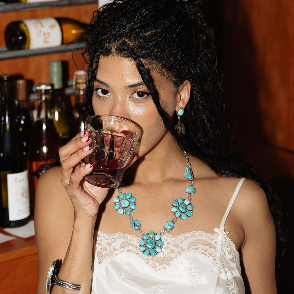 Woman in a white dress drinking from a glass with bottles in the background