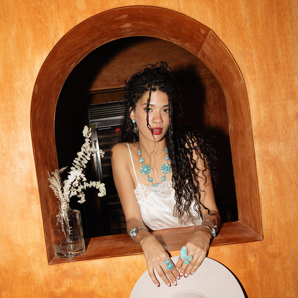Woman sitting inside a wooden alcove with a white hat, wearing a white dress and blue jewelry.