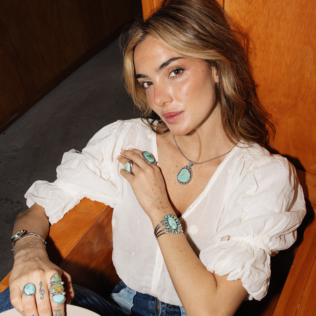 Woman wearing a white blouse and blue jeans with jewelry, sitting against a wooden wall.