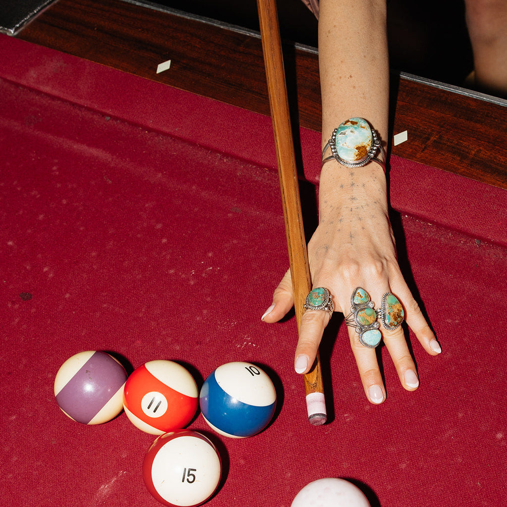 Hand with colorful rings reaching towards pool balls on a red felt table.