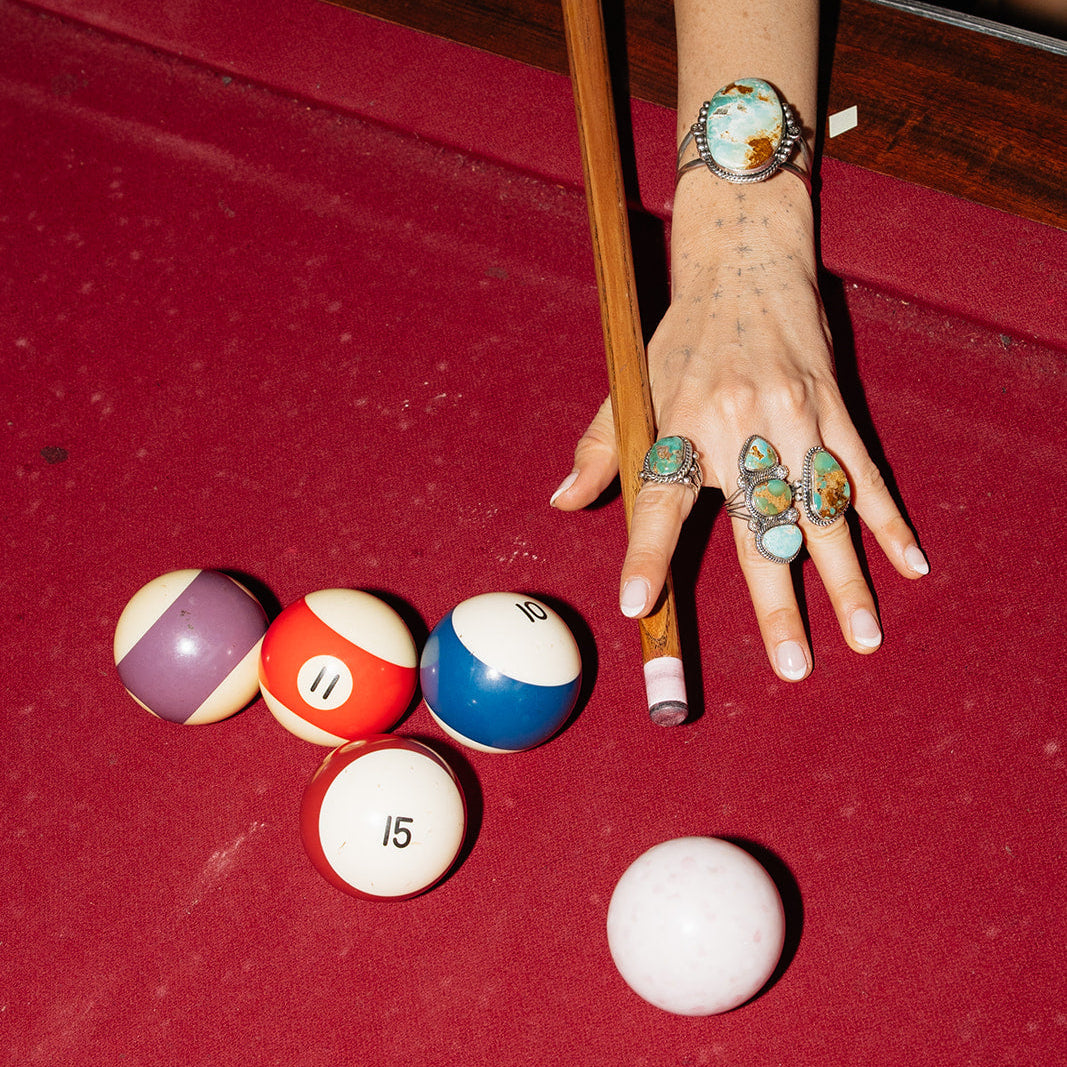 Hand with colorful rings reaching towards pool balls on a red felt table.