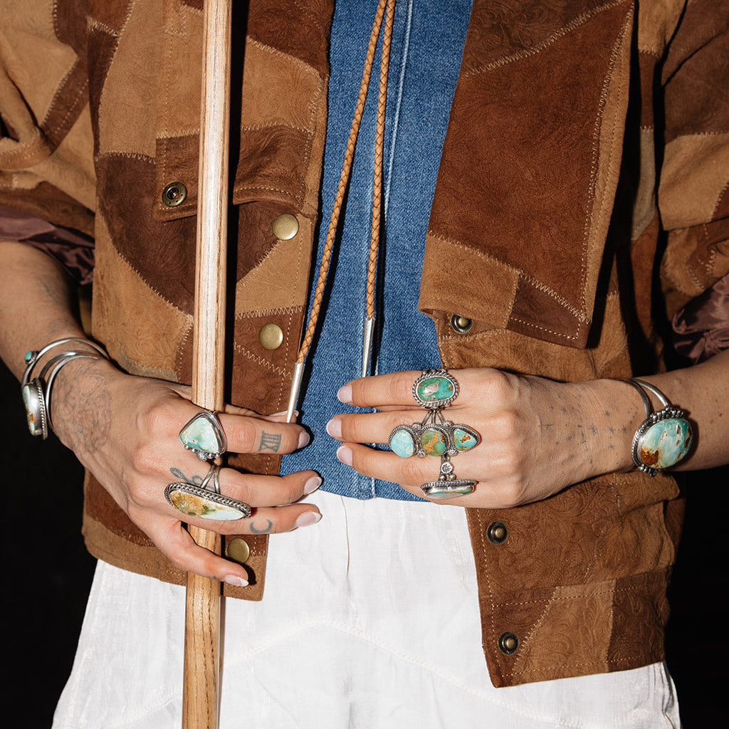 Person wearing a brown patterned jacket with jewelry and holding a wooden object.