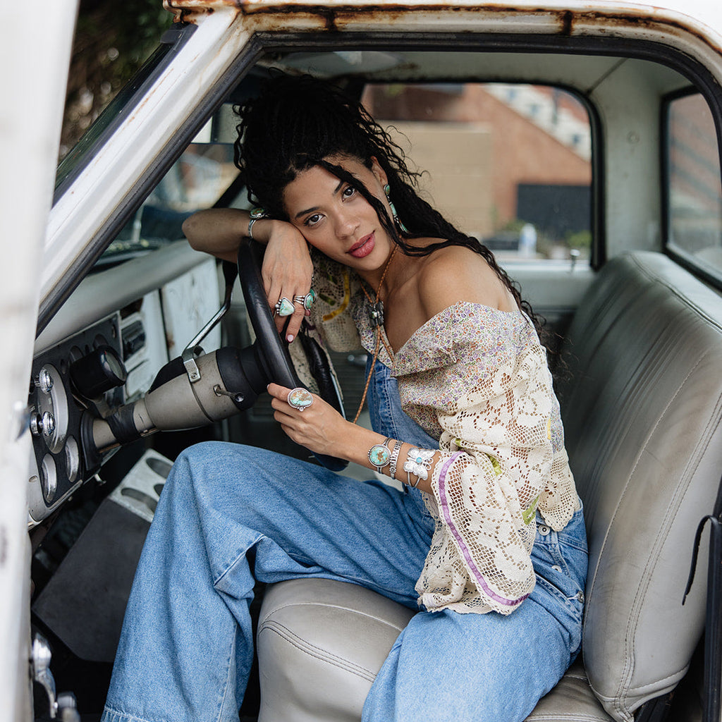 Woman sitting inside a vintage car wearing a patterned top and blue jeans.