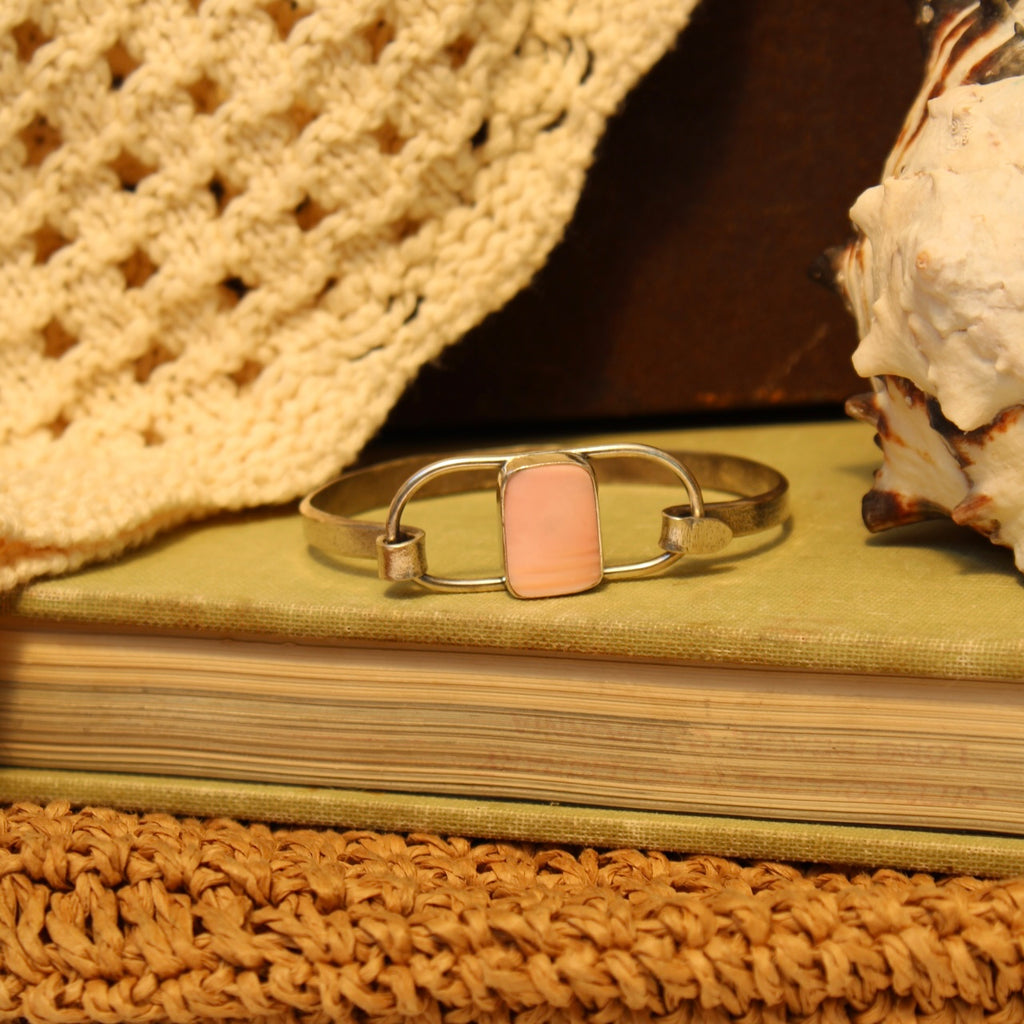A silver-toned metal cuff bracelet with a rectangular light pink stone, displayed on a vintage book with a crochet hat, seashell, and woven mat in the background.