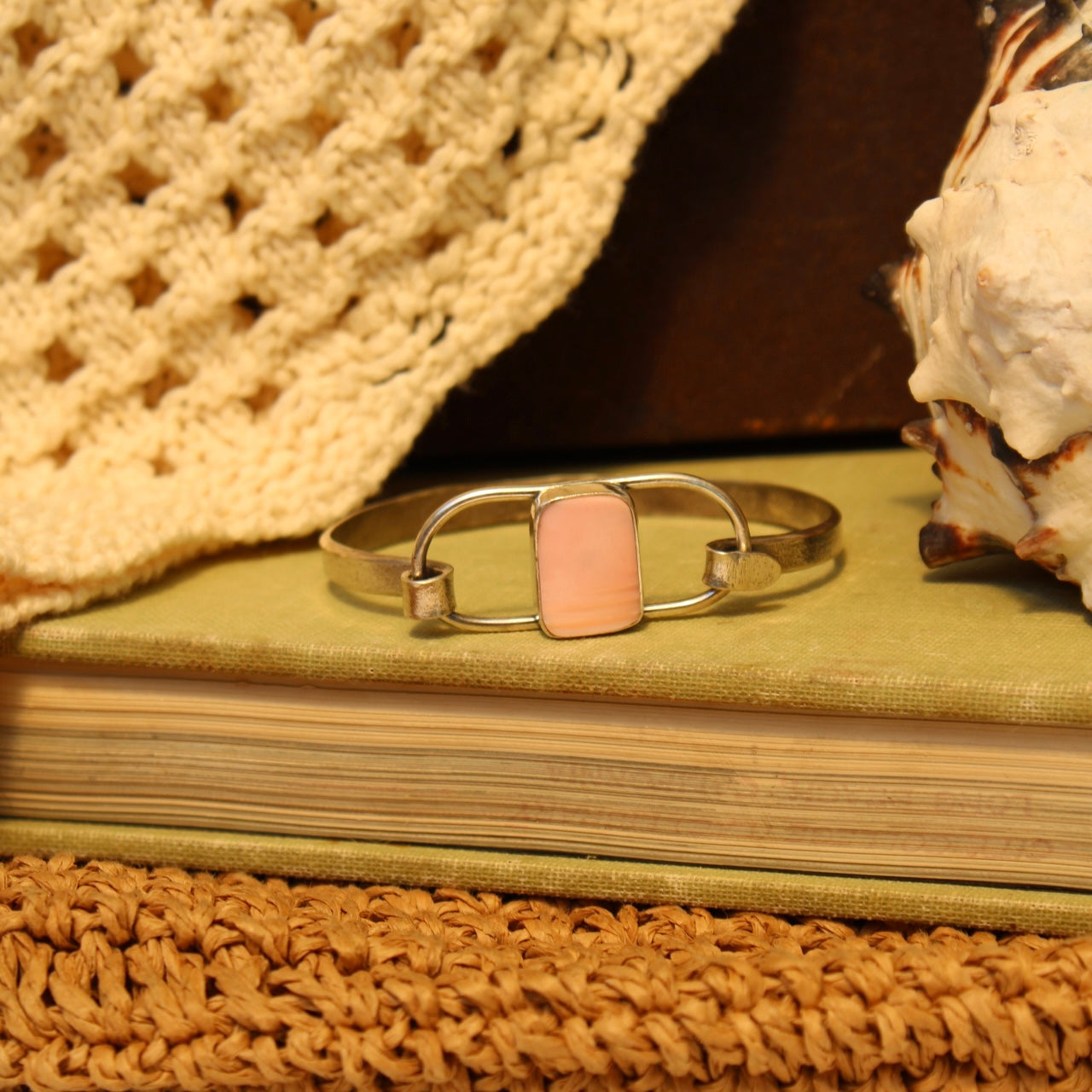 A silver-toned metal cuff bracelet with a rectangular light pink stone, displayed on a vintage book with a crochet hat, seashell, and woven mat in the background.