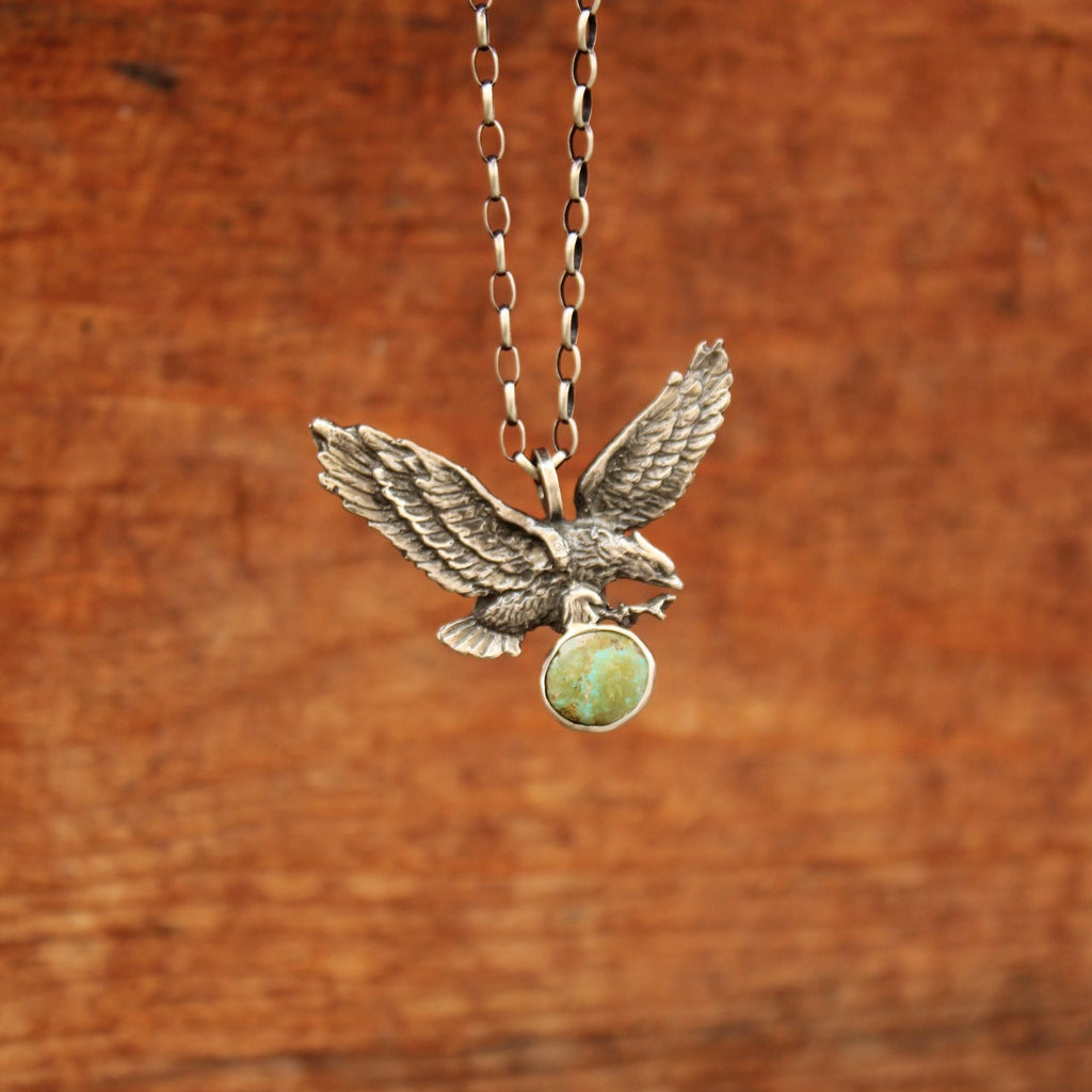 A silver-toned bird pendant necklace with a green oval stone, shown close-up against a warm brown wooden background.
