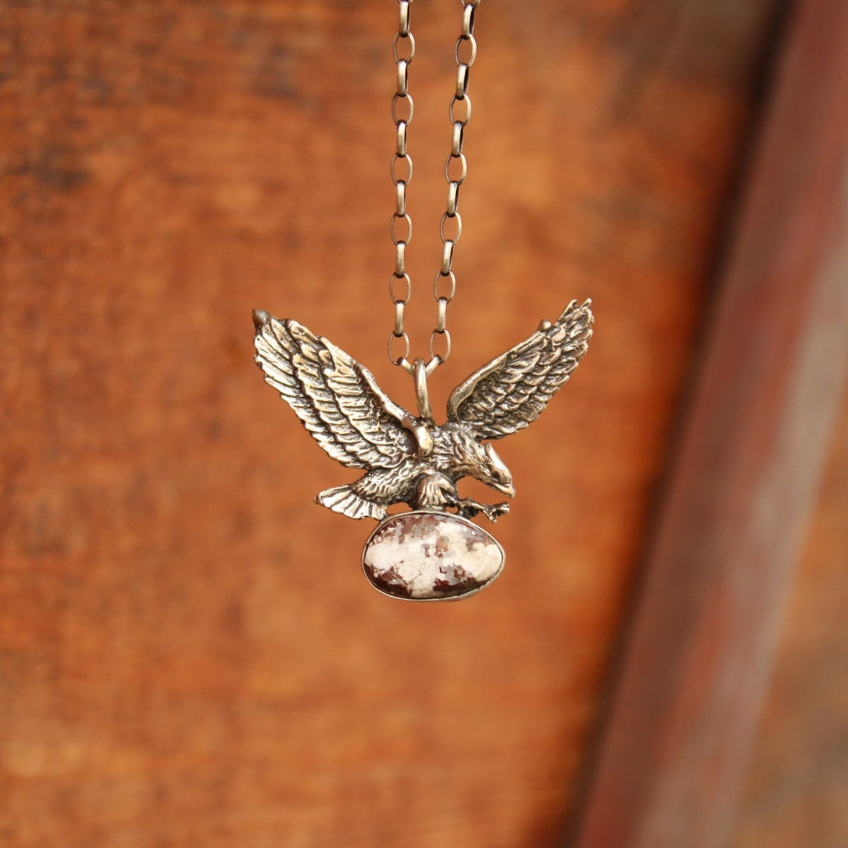 A silver-toned metal pendant necklace featuring an eagle holding a small oval stone, hanging from a matching chain against a warm brown wooden background.