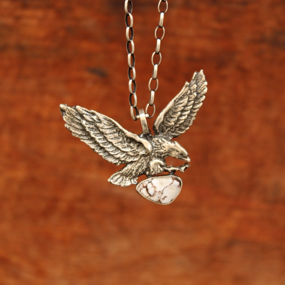 A close-up of a silver-toned pendant necklace featuring a detailed eagle holding a small white stone, suspended from a matching chain against a warm brown textured background.