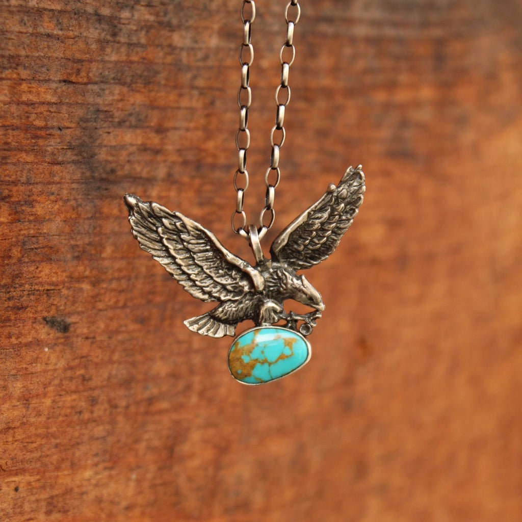 An antiqued silver eagle pendant necklace holding a turquoise stone, hanging against a rustic wooden background.