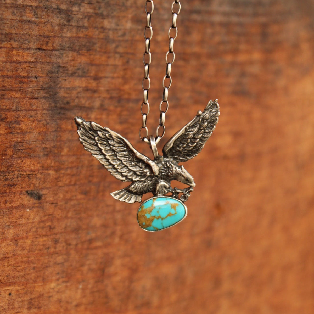 An antiqued silver eagle pendant necklace holding a turquoise stone, hanging against a rustic wooden background.