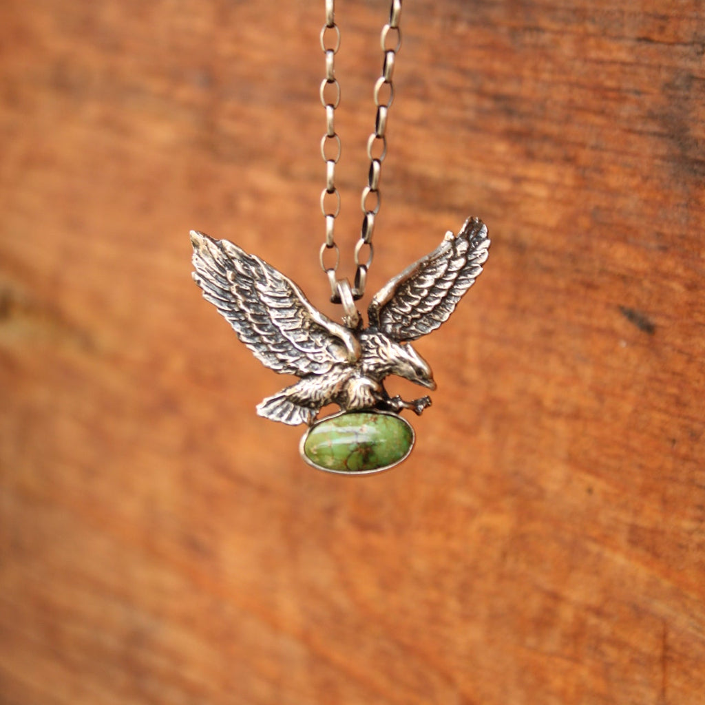 A silver-toned metal pendant shaped like a flying bird with spread wings and a smooth green oval stone, attached to a delicate chain against a warm brown wood background.