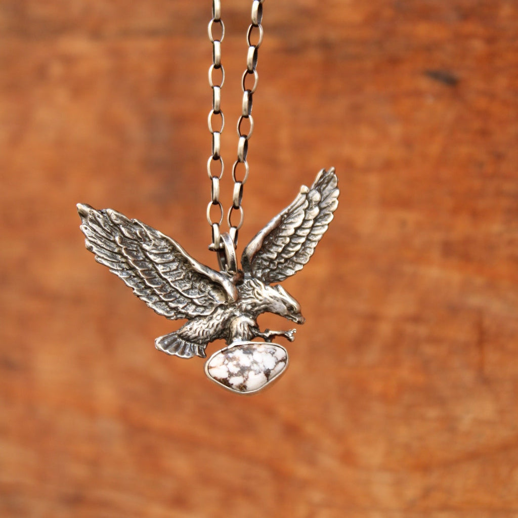 A silver-toned metal pendant shaped like a flying eagle clutching a shield, attached to a matching chain, shown against a warm brown wood-grain background.