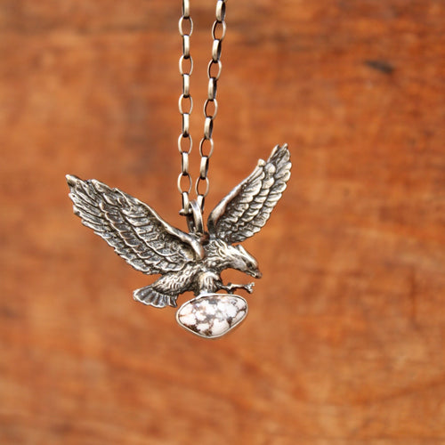 A silver-toned metal pendant shaped like a flying eagle clutching a shield, attached to a matching chain, shown against a warm brown wood-grain background.