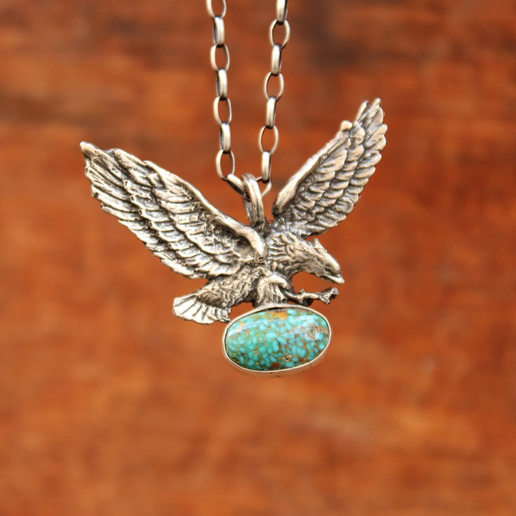 A silver-toned eagle pendant necklace with a textured, feathered body and a round turquoise-colored stone on its head, shown on a matching chain against a warm brown wooden background.