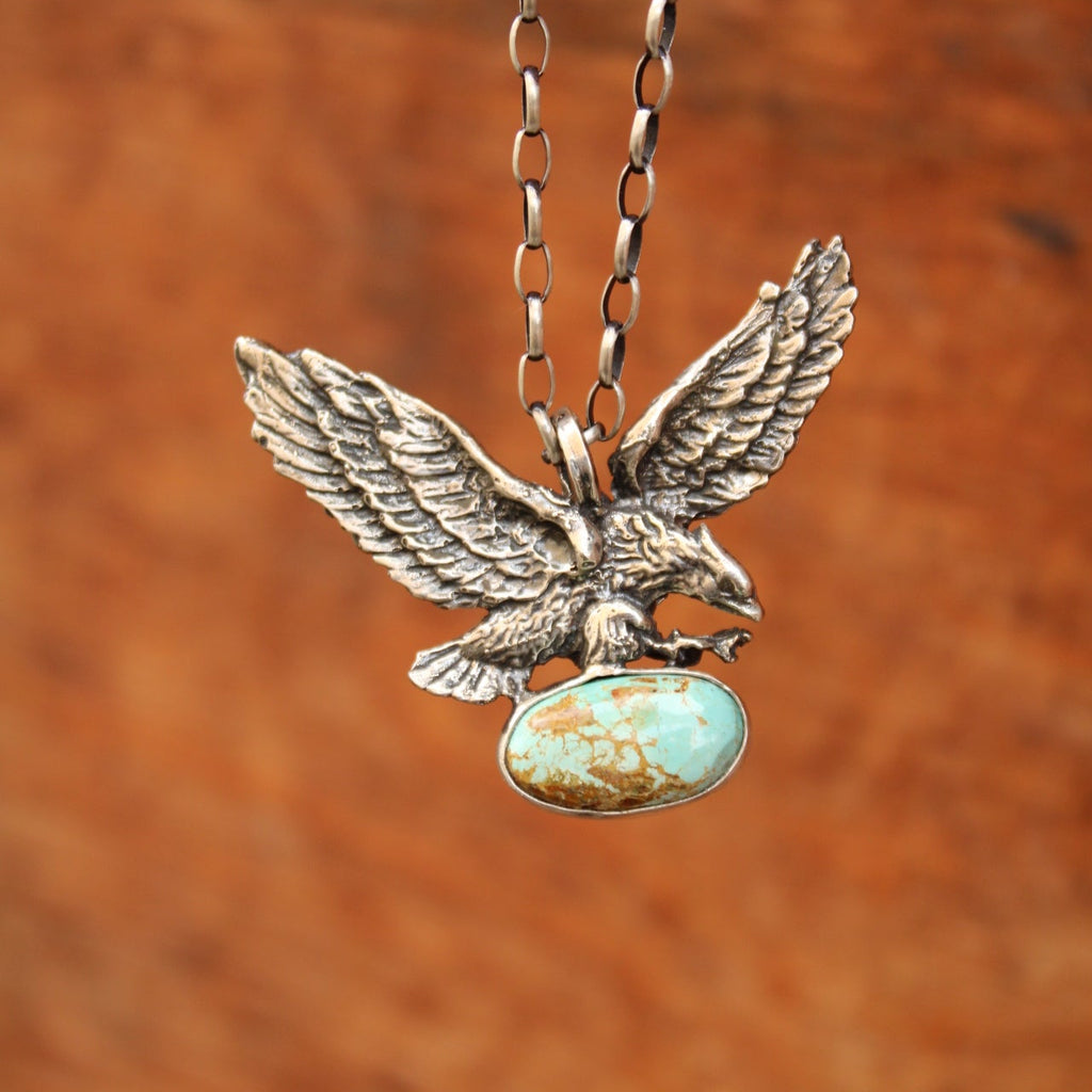 A close-up of a silver-toned eagle pendant necklace with a turquoise-colored oval stone, shown against a warm brown textured background.