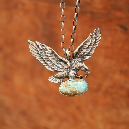 A close-up of a silver-toned eagle pendant necklace with a turquoise-colored oval stone, shown against a warm brown textured background.