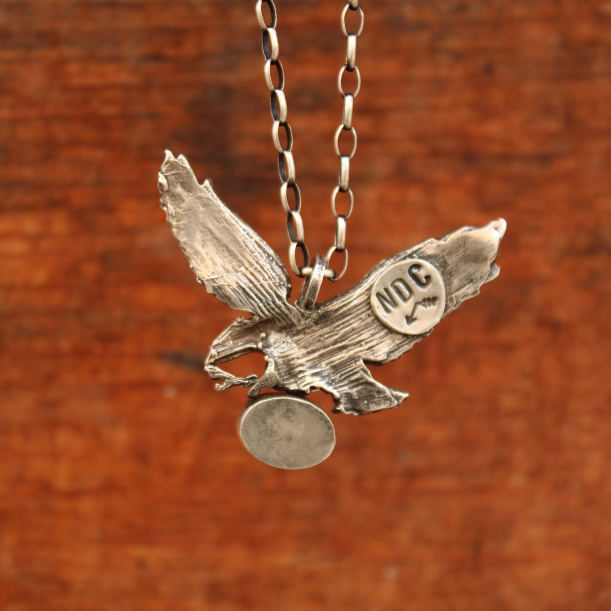 A sterling silver pendant necklace featuring a textured bird design with a circular medallion engraved with '302' on its chest, suspended from a silver link chain against a warm, rustic brown wooden background.