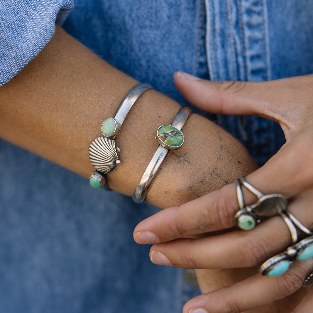 Close-up of a person's wrist and hand wearing silver bracelets and rings with green stones against a blue denim background.