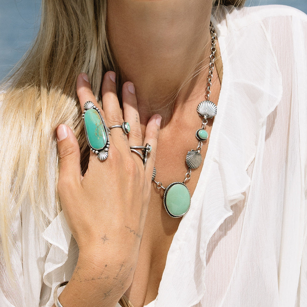 Woman wearing turquoise jewelry including rings, a necklace, and earrings by a waterfront.