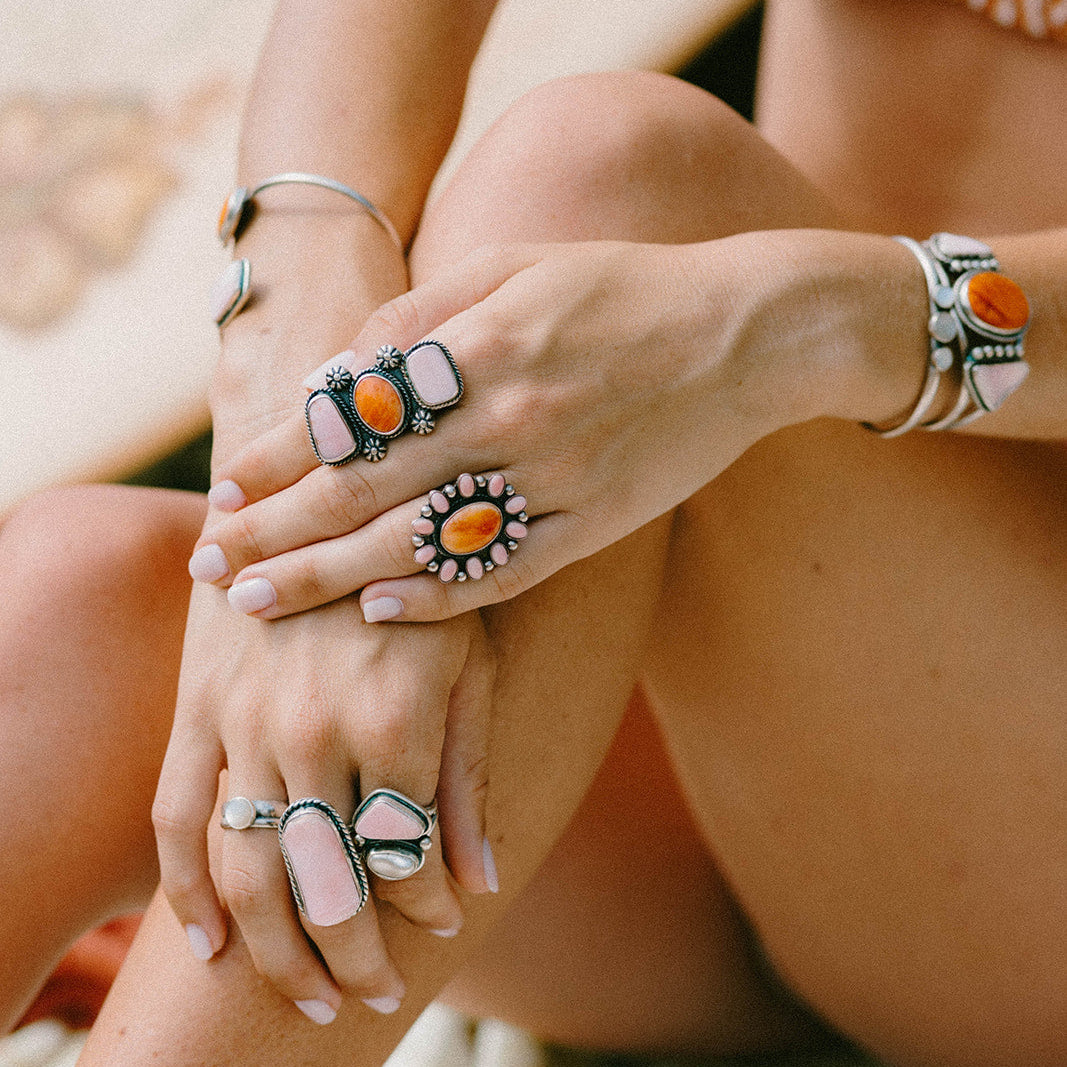 A close-up lifestyle shot of a person's hands and legs wearing multiple bohemian-style rings and bracelets with orange, pink, and white stones on a neutral background.