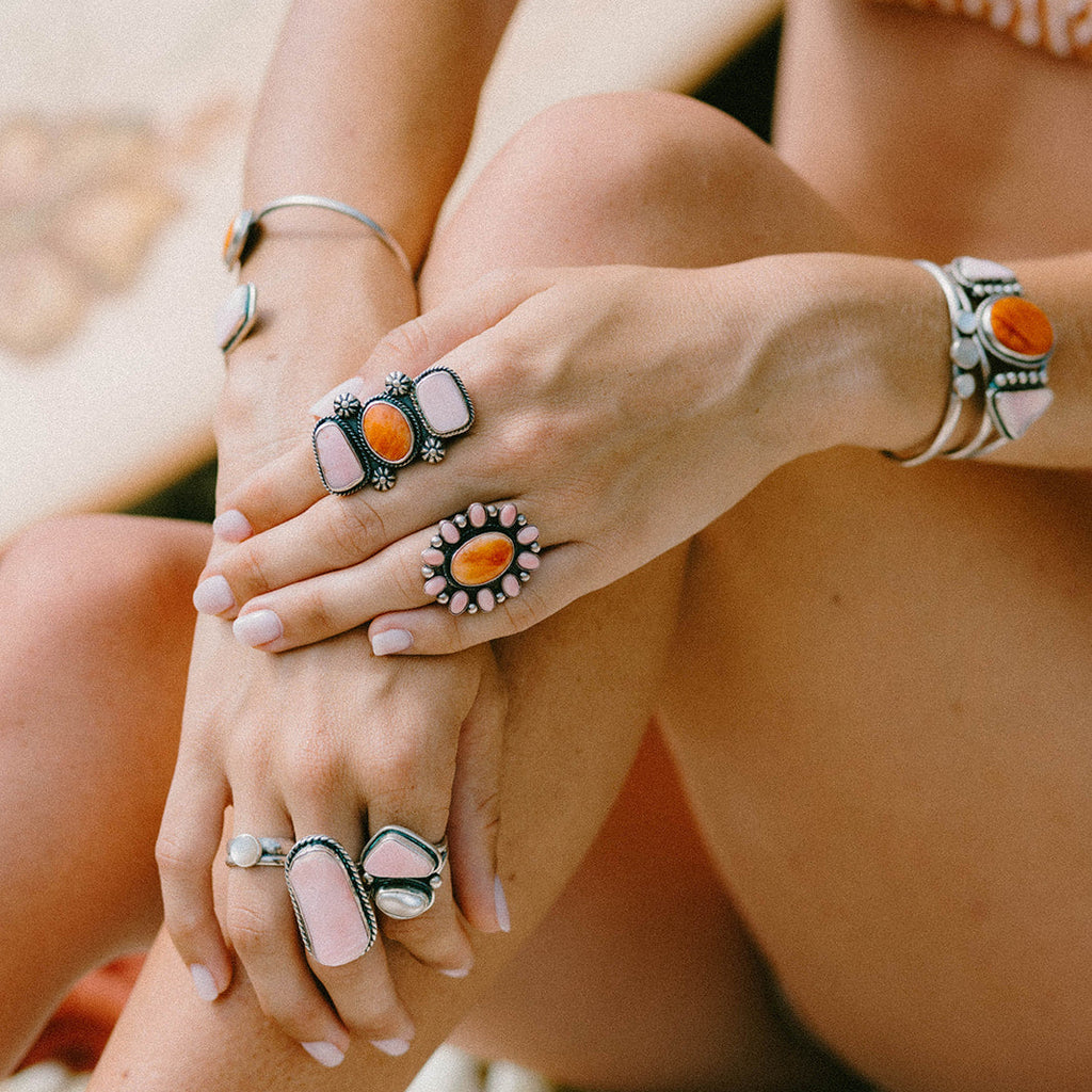A close-up lifestyle shot of a person's hands and legs wearing multiple bohemian-style rings and bracelets with orange, pink, and white stones on a neutral background.