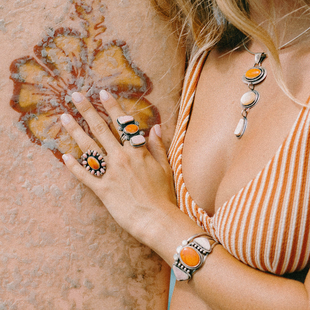 A woman wearing a matching set of silver-toned jewelry with orange and white accents, including a necklace, bracelet, and rings, posing against a textured stucco wall.