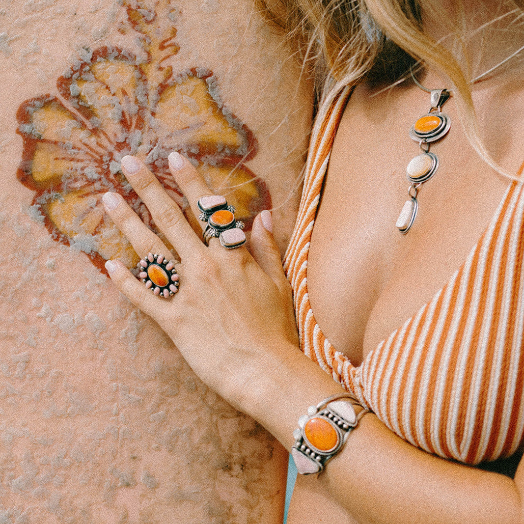 A woman wearing a matching set of silver-toned jewelry with orange and white accents, including a necklace, bracelet, and rings, posing against a textured stucco wall.