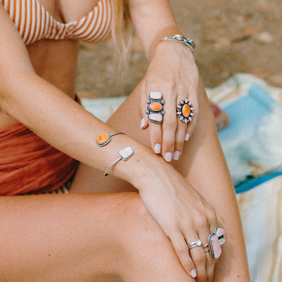 A woman wearing multiple silver-toned rings with orange and white stone details, along with bracelets, in a beach setting.