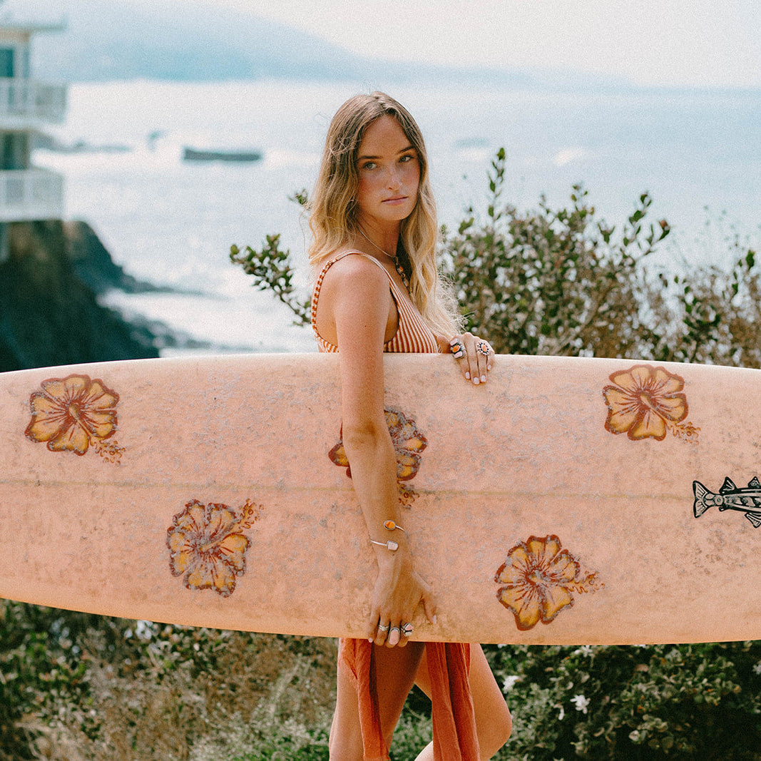 A woman stands on a cliffside overlooking the ocean, holding a light pink surfboard decorated with orange hibiscus flowers and a fish graphic. A large white coastal building is visible in the background.