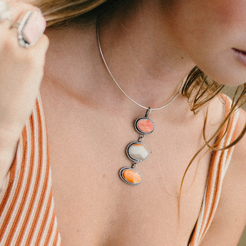 A close-up lifestyle shot of a woman wearing a delicate silver chain necklace with three vertically stacked oval charms in coral pink, cream, and orange, paired with an orange and white striped top.
