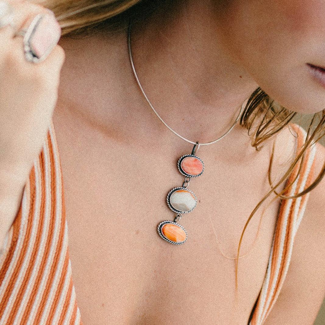 A close-up lifestyle shot of a woman wearing a delicate silver chain necklace with three vertically stacked oval charms in coral pink, cream, and orange, paired with an orange and white striped top.