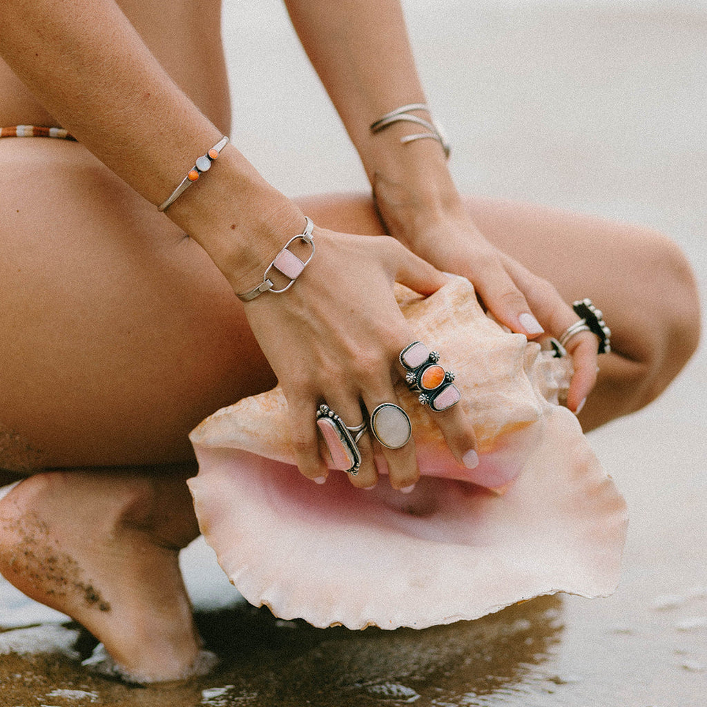 A close-up lifestyle shot of a person crouching on a sandy beach with their hands resting on a large pink seashell. The person is wearing multiple silver-toned rings with colorful stones and several silver bracelets.