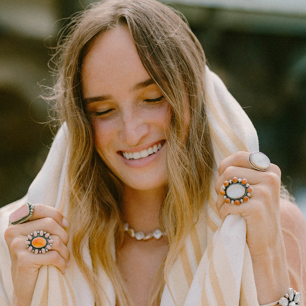 A woman with long blonde hair smiling while wrapped in a cream and tan striped towel, wearing multiple bohemian-style rings and bracelets.