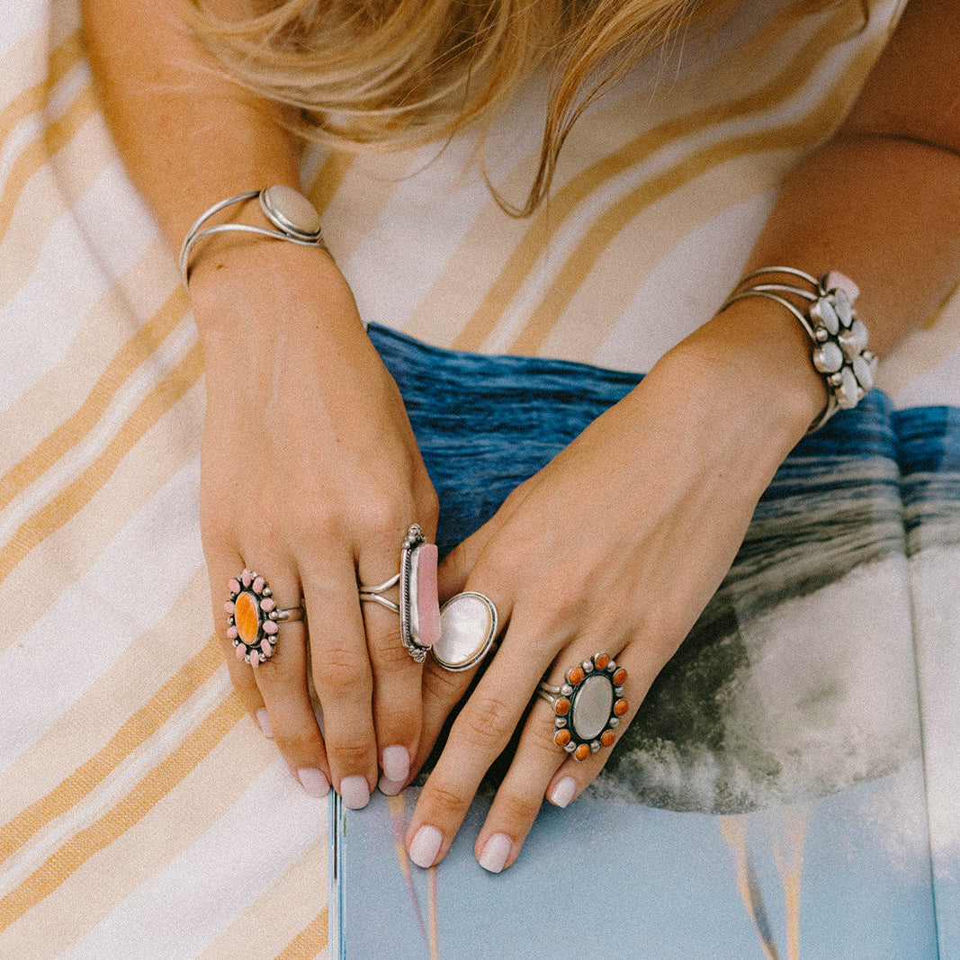 A close-up lifestyle shot of a person's hands and arms adorned with multiple silver-toned rings and bracelets, resting on a striped blanket over a blue and white abstract art print.