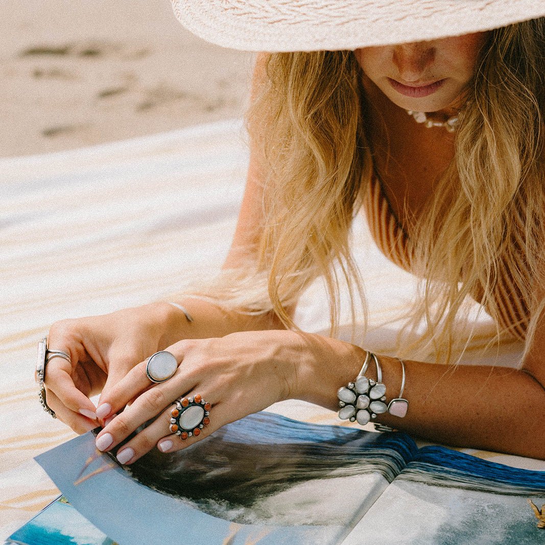 A woman wearing a large woven straw hat and bohemian jewelry lies on a striped beach towel at the beach, looking down at an open book or magazine.