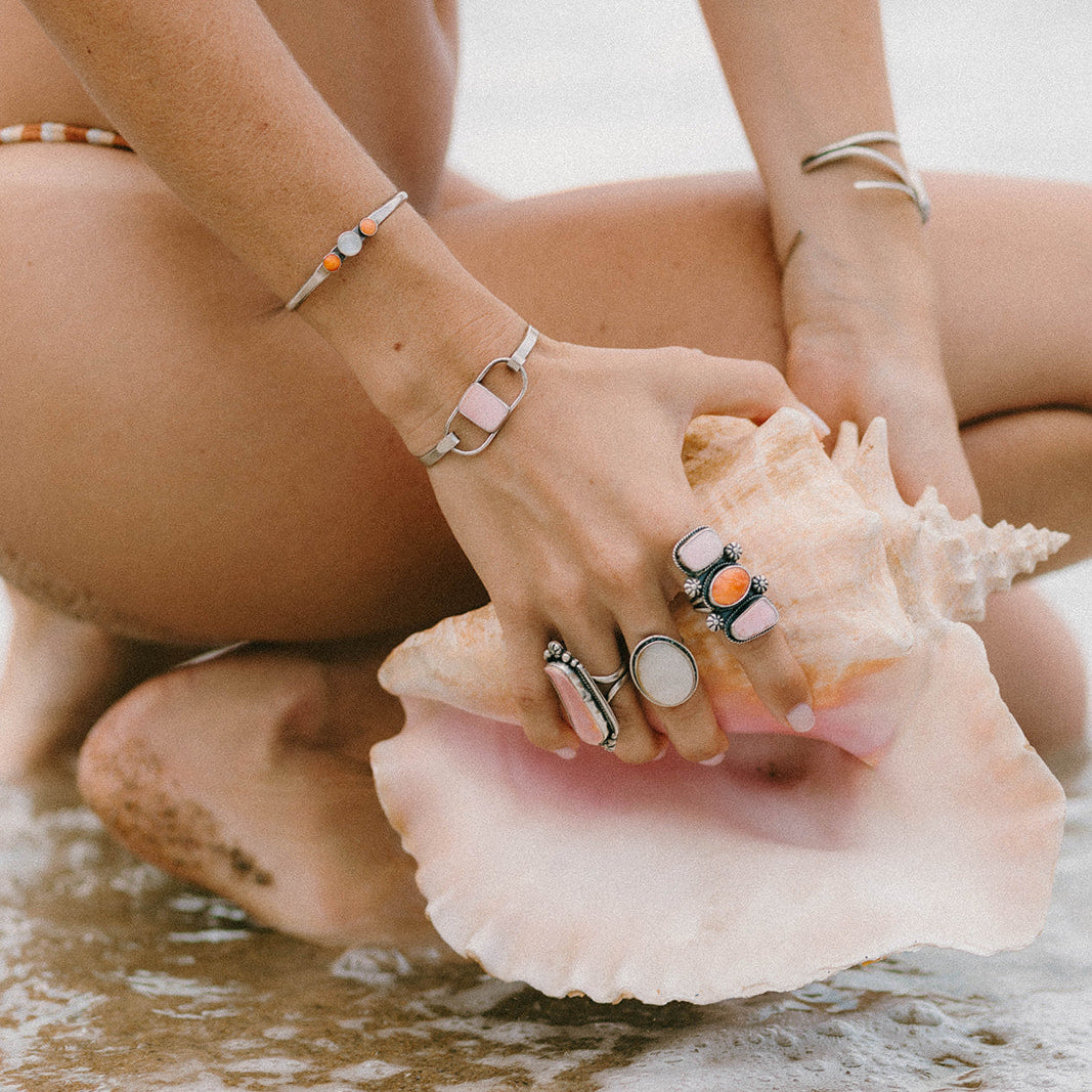 A close-up lifestyle shot of a person sitting on wet sand at the beach, holding a large seashell. The person is wearing multiple silver-toned rings and bracelets, including one prominent ring with a white oval stone and another with a black and orange graphic.