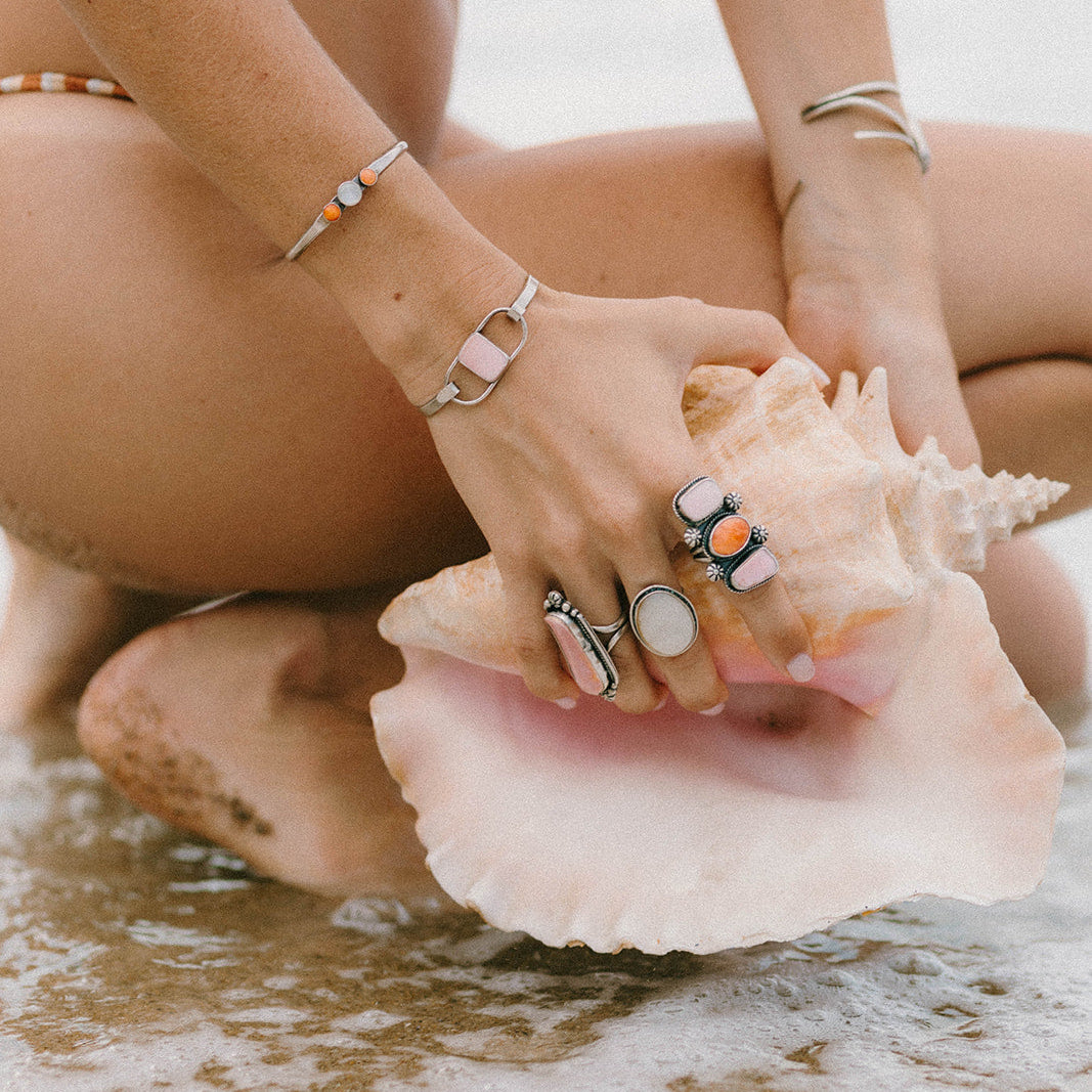 A close-up lifestyle shot of a person sitting on wet sand at the beach, holding a large seashell. The person is wearing multiple silver-toned rings and bracelets, including one prominent ring with a white oval stone and another with a black and orange graphic.