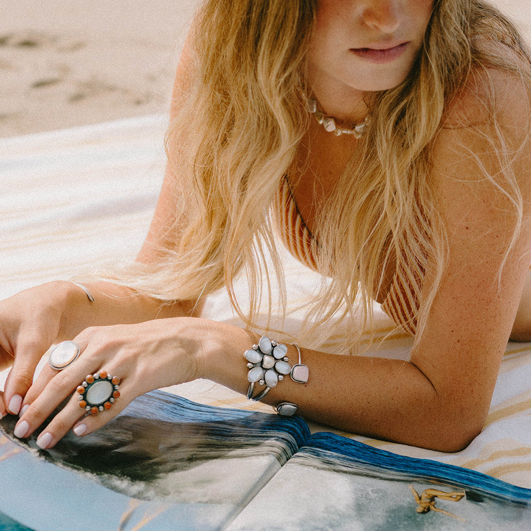 A woman reclining on a beach towel at the sand, wearing a wide-brimmed straw hat, a pearl necklace, and multiple silver-toned rings and bracelets, with an open blue and white book in front of her.