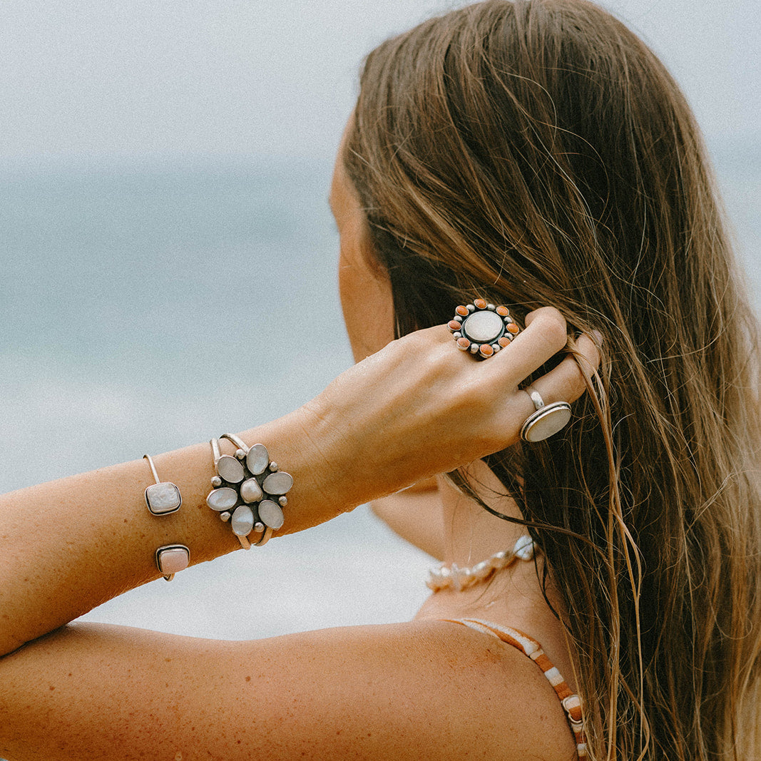 A woman with long brown hair seen from the back and side, wearing multiple bohemian-style jewelry pieces including a flower-shaped cuff bracelet, a silver-toned bangle, a ring, and a necklace, with a beach background.